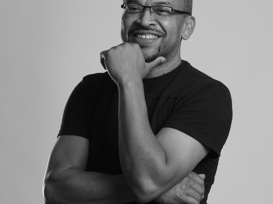 A black mid aged man stands with his arms crossed and chin resting in hand, smiling